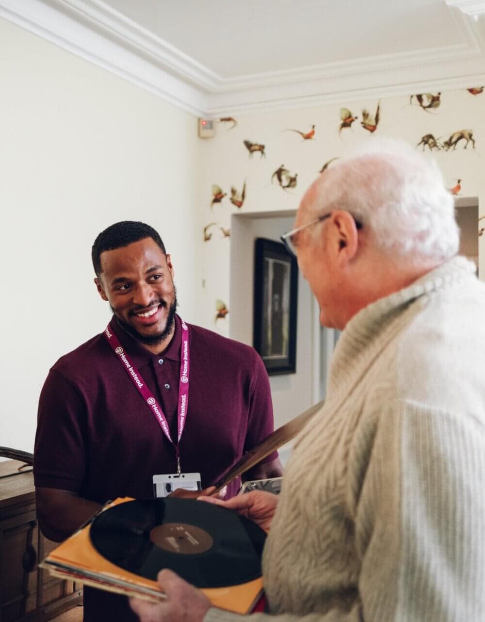 A smiling man in a maroon shirt and lanyard talks to an older man holding a vinyl record in a cozy, decorated room. - Home Instead