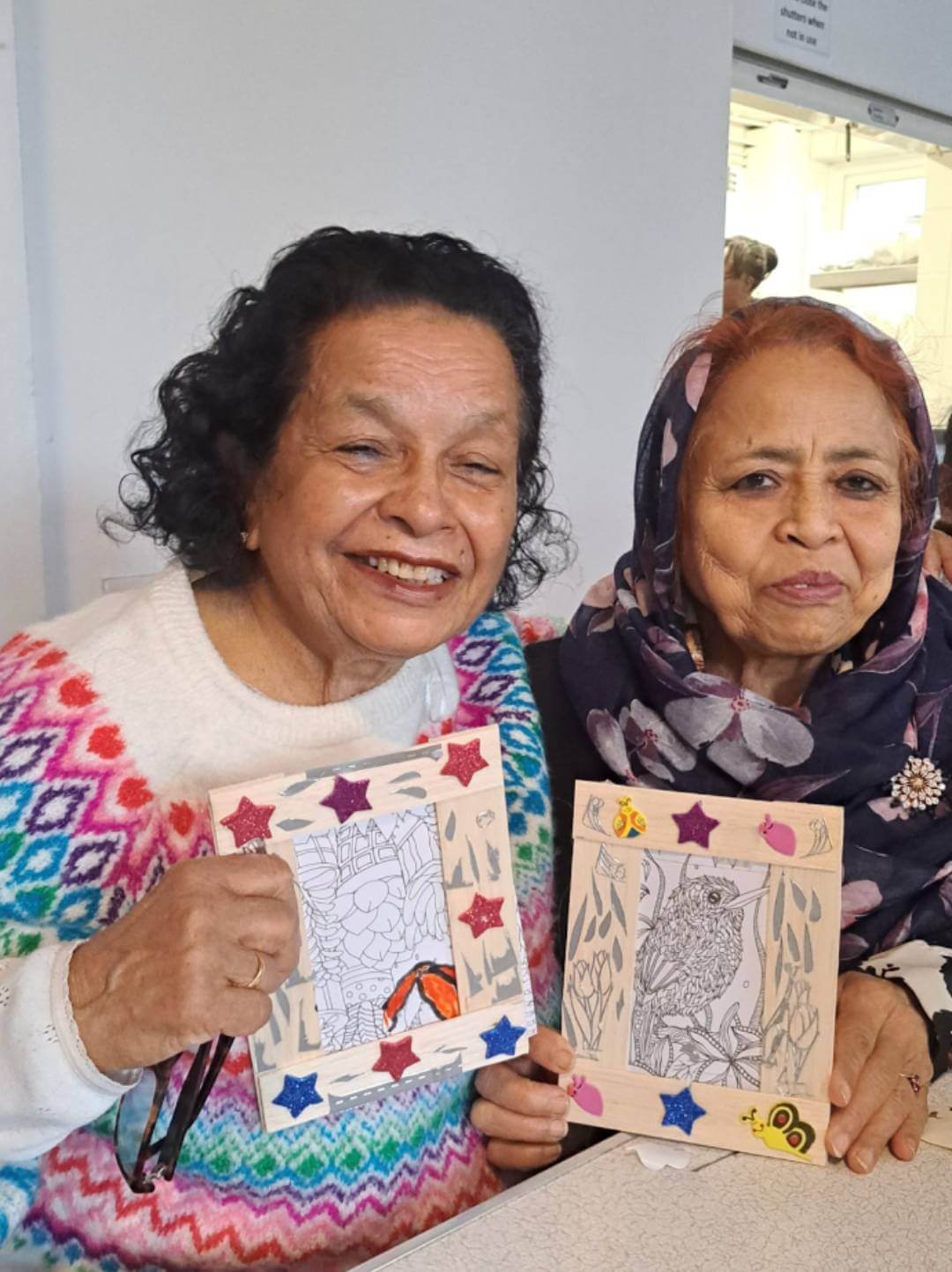 Two smiling elderly women holding decorated picture frames with intricate drawings, sharing a joyful moment together. - Home Instead