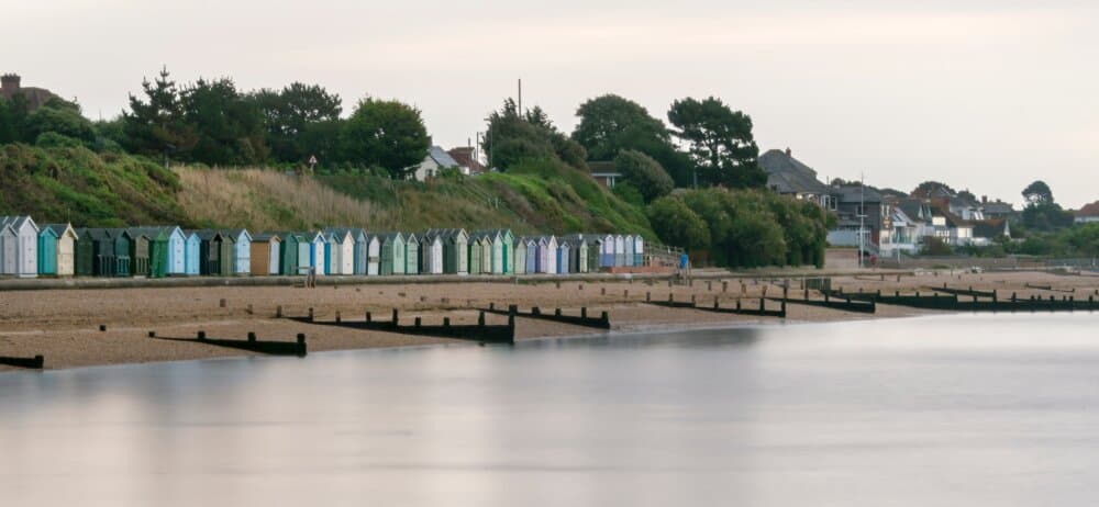 A row of colorful beach huts on a pebble beachfront with calm water in the foreground and trees and houses in the background. - Home Instead