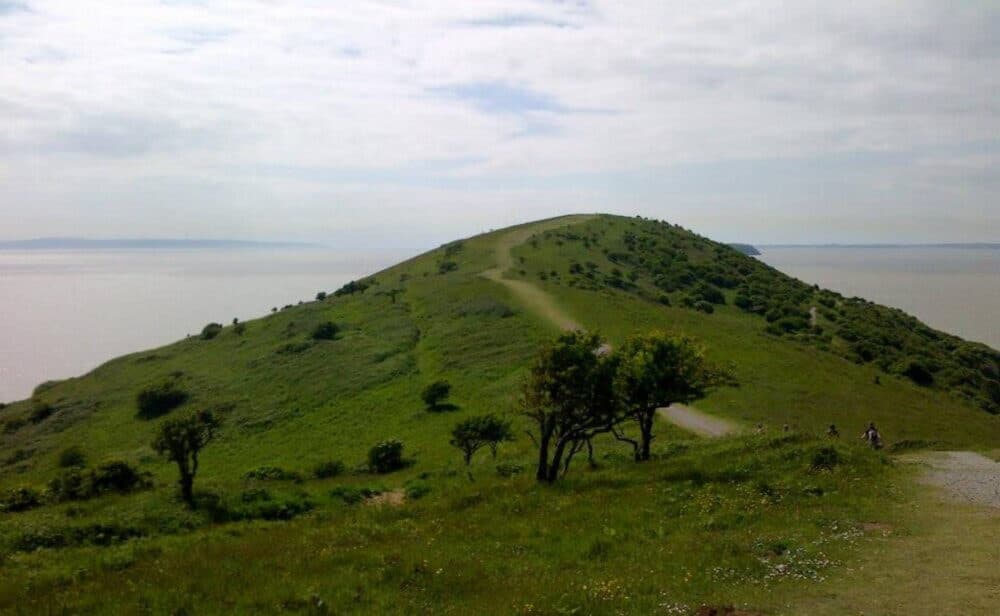 A grassy hill with a footpath winding along the top, leading towards the coastline under a partly cloudy sky. - Home Instead