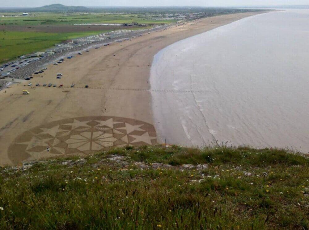 Beach with large star and geometric pattern drawn in the sand, view from a high cliff overlooking the coast. - Home Instead