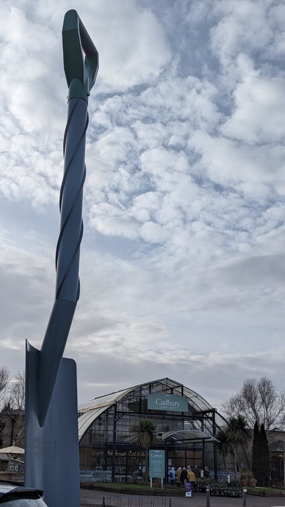 Tall twisted metal sculpture in front of the Cadbury World building on a cloudy day with some blue sky patches. - Home Instead