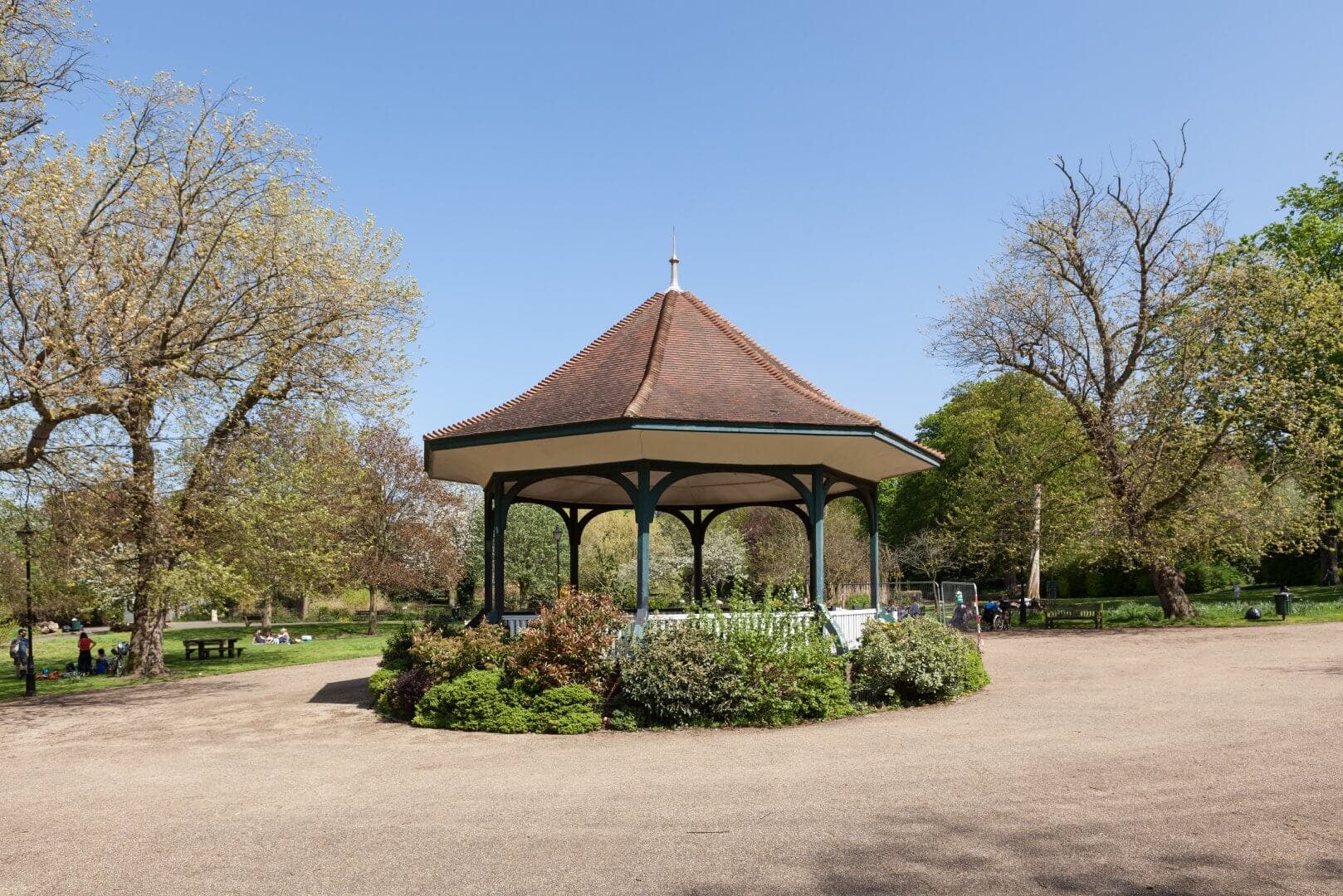 A gazebo with a red roof in a park surrounded by greenery on a sunny day with a clear blue sky. - Home Instead