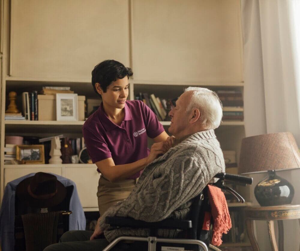 A caregiver helps an elderly man in a wheelchair with his sweater in a cozy, book-filled room. - Home Instead
