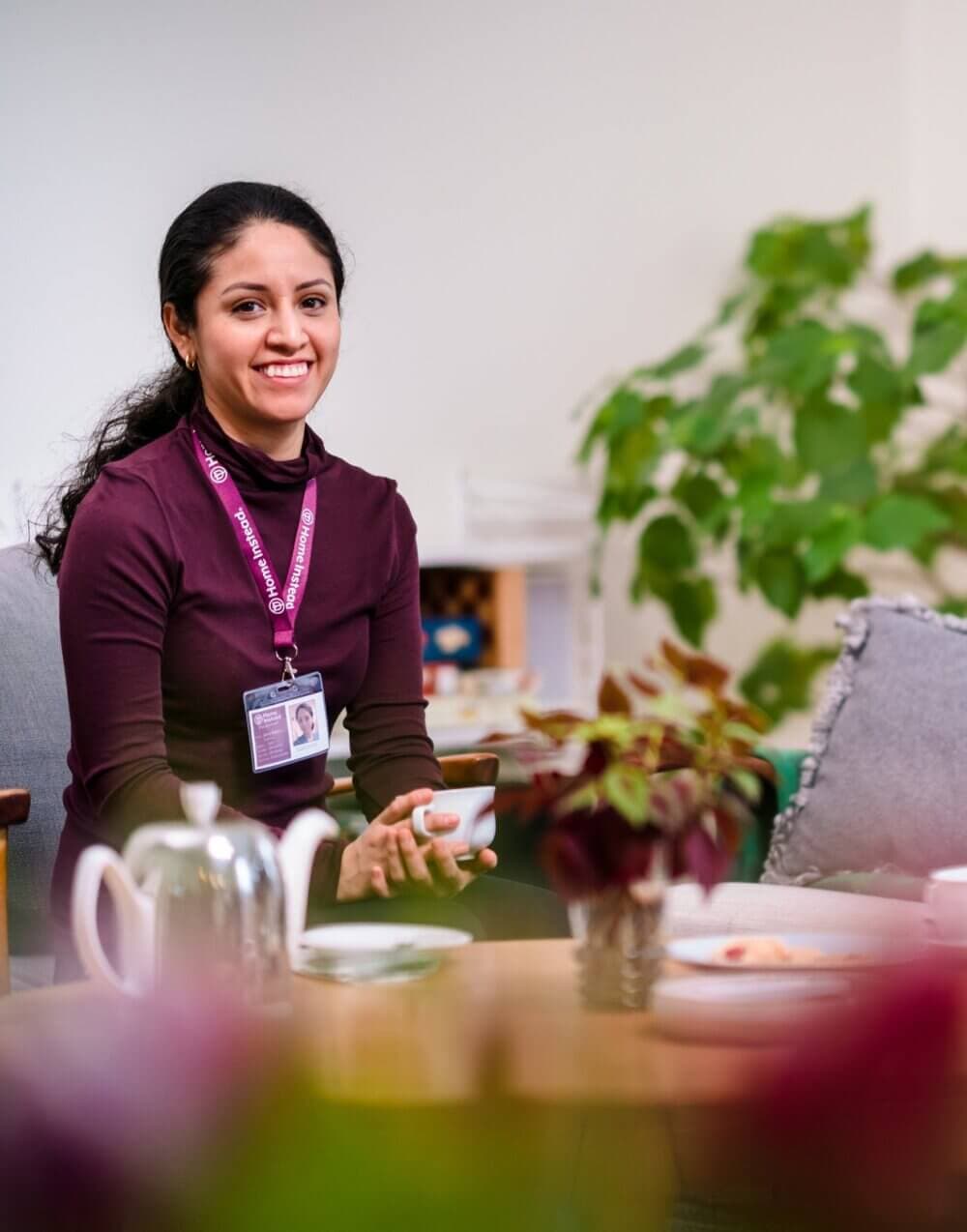 Smiling woman with ID badge holding cup, seated at a table with tea set and plant in the background. - Home Instead