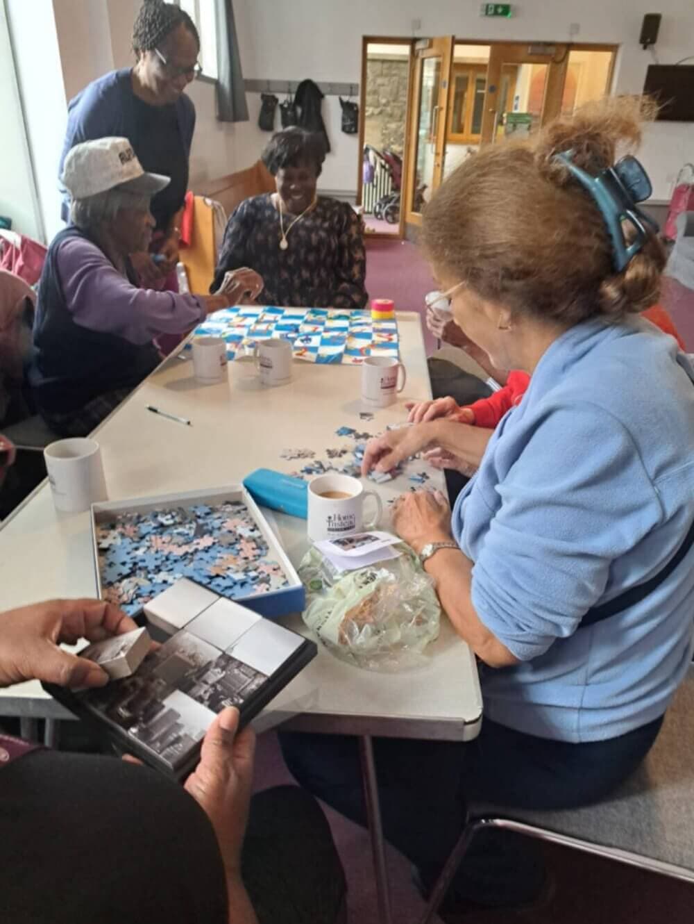 A group of people sitting at a table doing puzzles and chatting in a community center room. - Home Instead