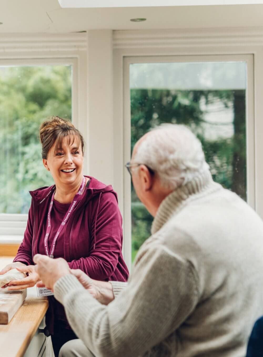 A woman and an elderly man seated at a table, smiling and engaging in conversation by a large window. - Home Instead