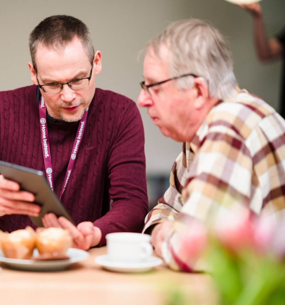 Two men sitting at a table, one showing the other a tablet. Cupcakes and coffee on the table. - Home Instead