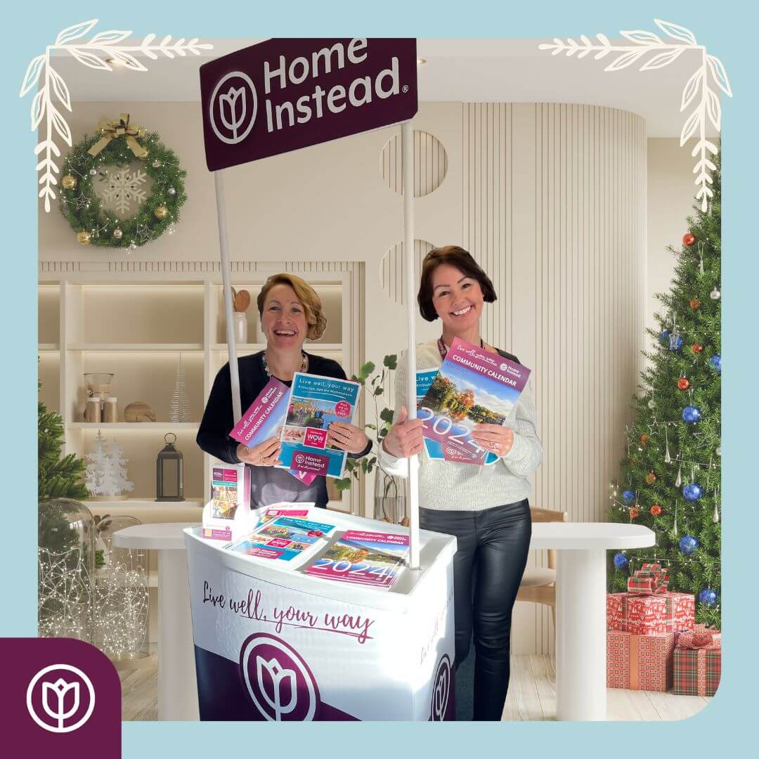 Two women at a Home Instead booth holding brochures. Christmas decorations and gifts are in the background. - Home Instead
