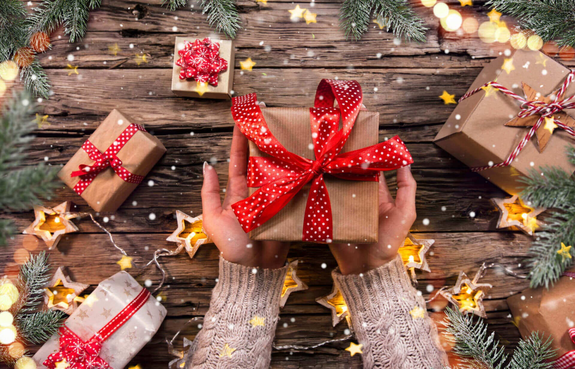 Hands holding a wrapped gift with a red ribbon, surrounded by other presents and festive decorations on a wooden surface. - Home Instead