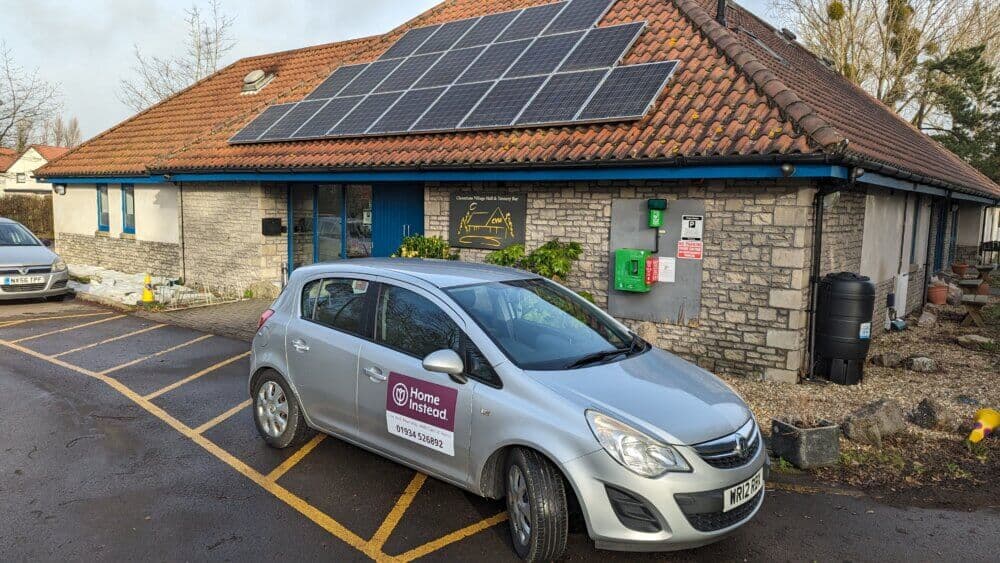 A silver car with "Home Instead" branding is parked in front of a building with solar panels on its roof. - Home Instead