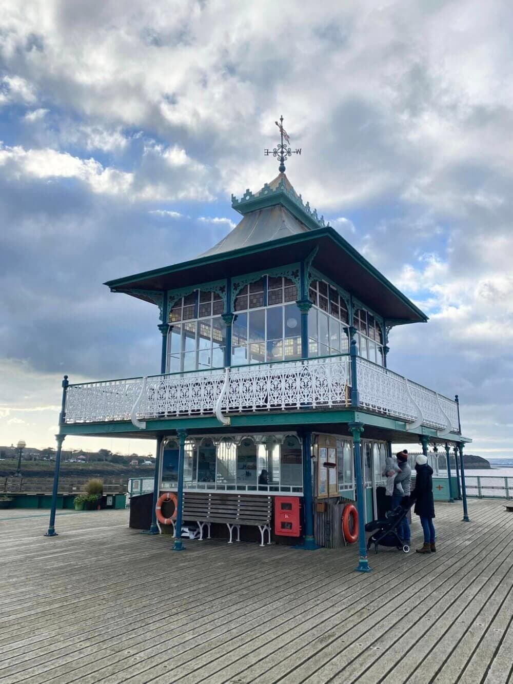 A couple with a stroller stands near a vintage pavilion on a pier, under a cloudy sky. - Home Instead