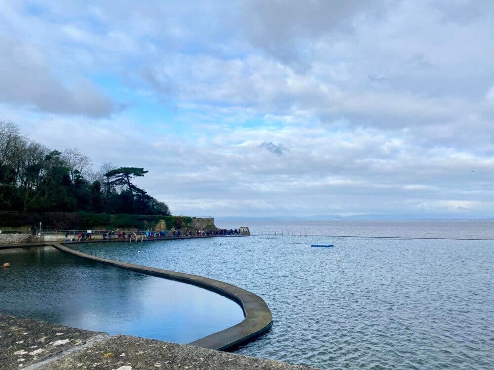 Outdoor swimming pool beside a calm sea, with a cloudy sky and a tree-lined shore in the background. - Home Instead