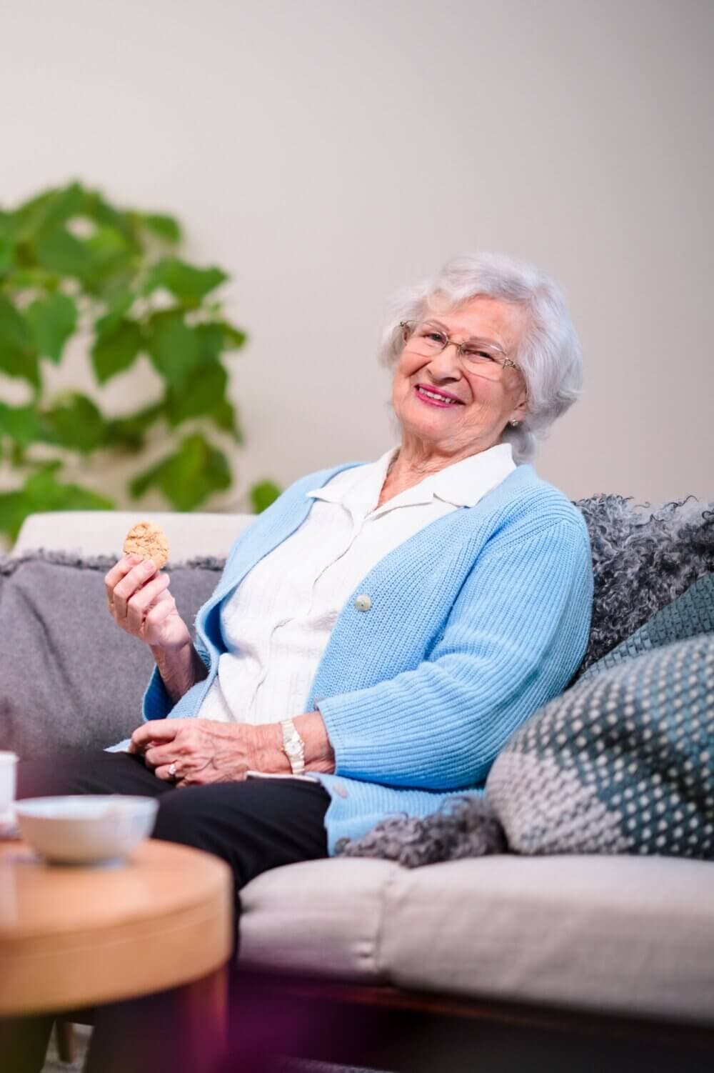 Elderly woman sitting on a couch, smiling, and holding a cookie with a plant in the background. - Home Instead