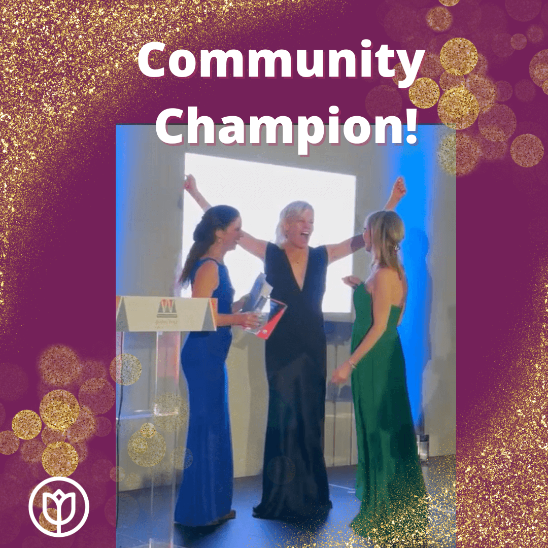 Three women in formal dresses celebrate on stage under a "Community Champion!" banner with glittery gold decorations. - Home Instead