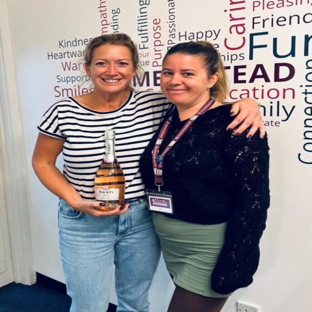 Two women smiling, one holding a bottle of sparkling wine, standing in front of a word-decorated wall. - Home Instead