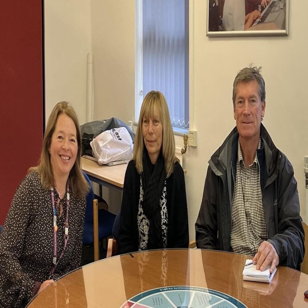 Three people seated around a wooden table in a conference room, smiling and posing for a photo. - Home Instead