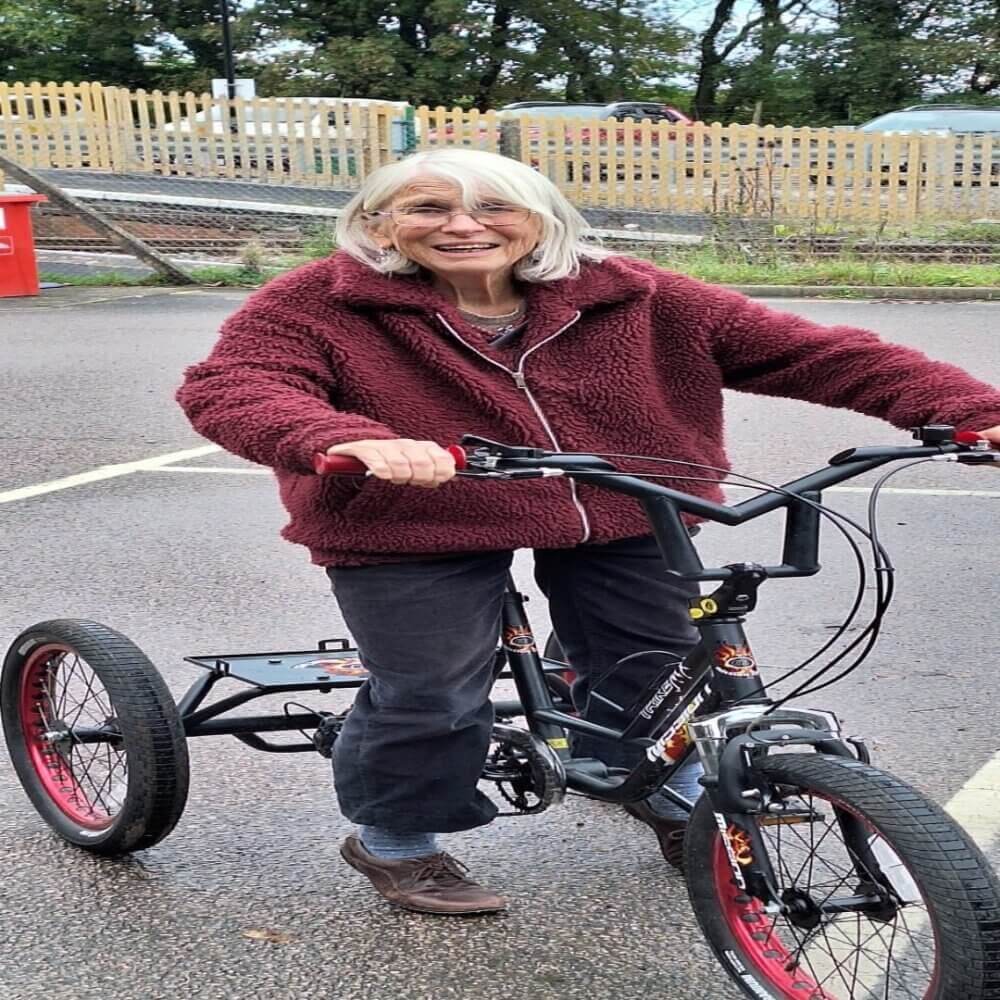 An elderly woman in a red jacket is smiling while standing with her hands on a three-wheeled bicycle. - Home Instead