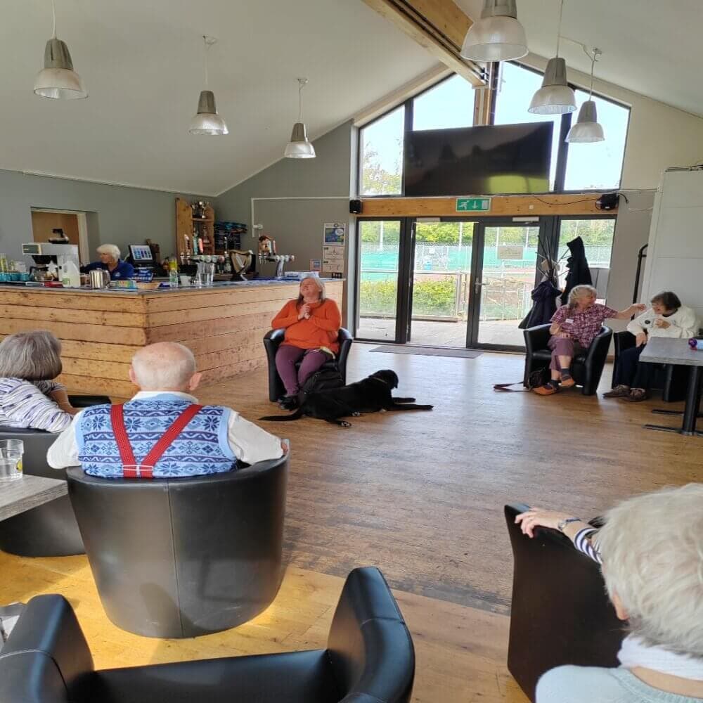 A group of seniors sitting in a cozy room with a woman in the center attending to a black dog. - Home Instead