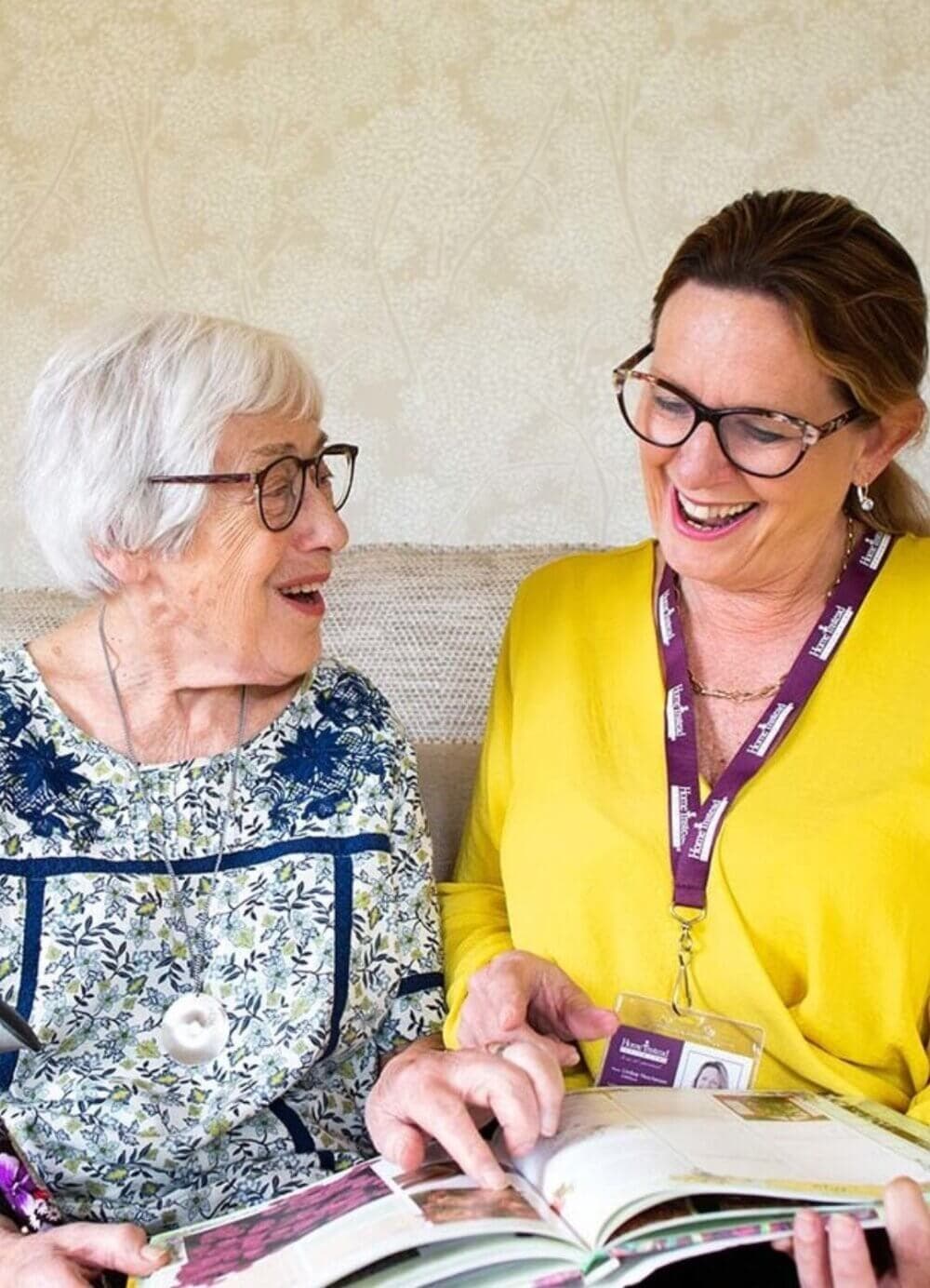Two women, one elderly and one younger, smiling and looking at a book together while sitting on a couch. - Home Instead