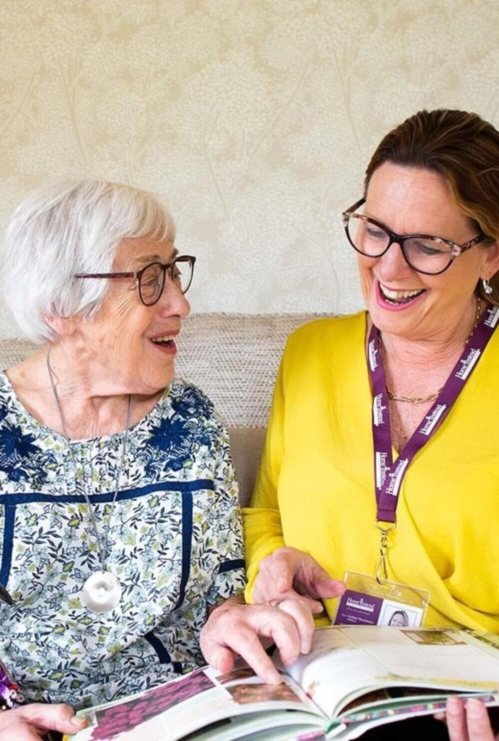 An elderly woman and a caregiver with a lanyard smiling while looking at an open book together. - Home Instead