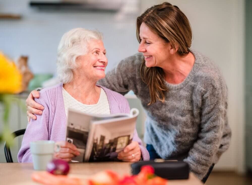 A smiling elderly woman reading a magazine is embraced by a younger woman in a cozy home setting. - Home Instead