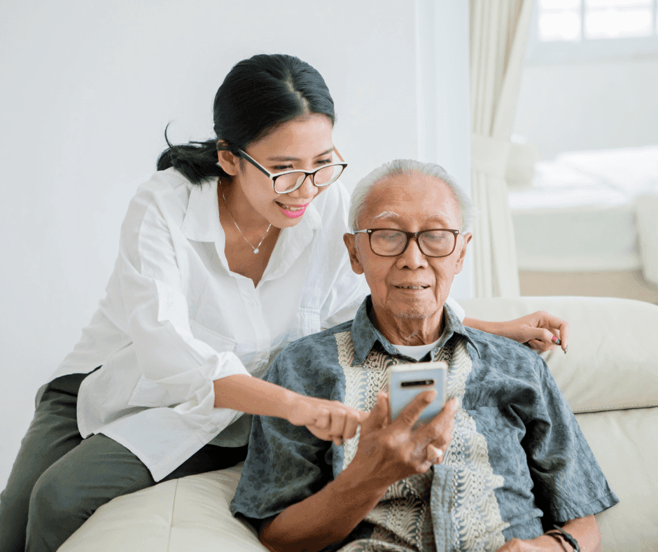 A young woman helps an elderly man with a smartphone. Both are smiling and sitting on a sofa in a bright room. - Home Instead