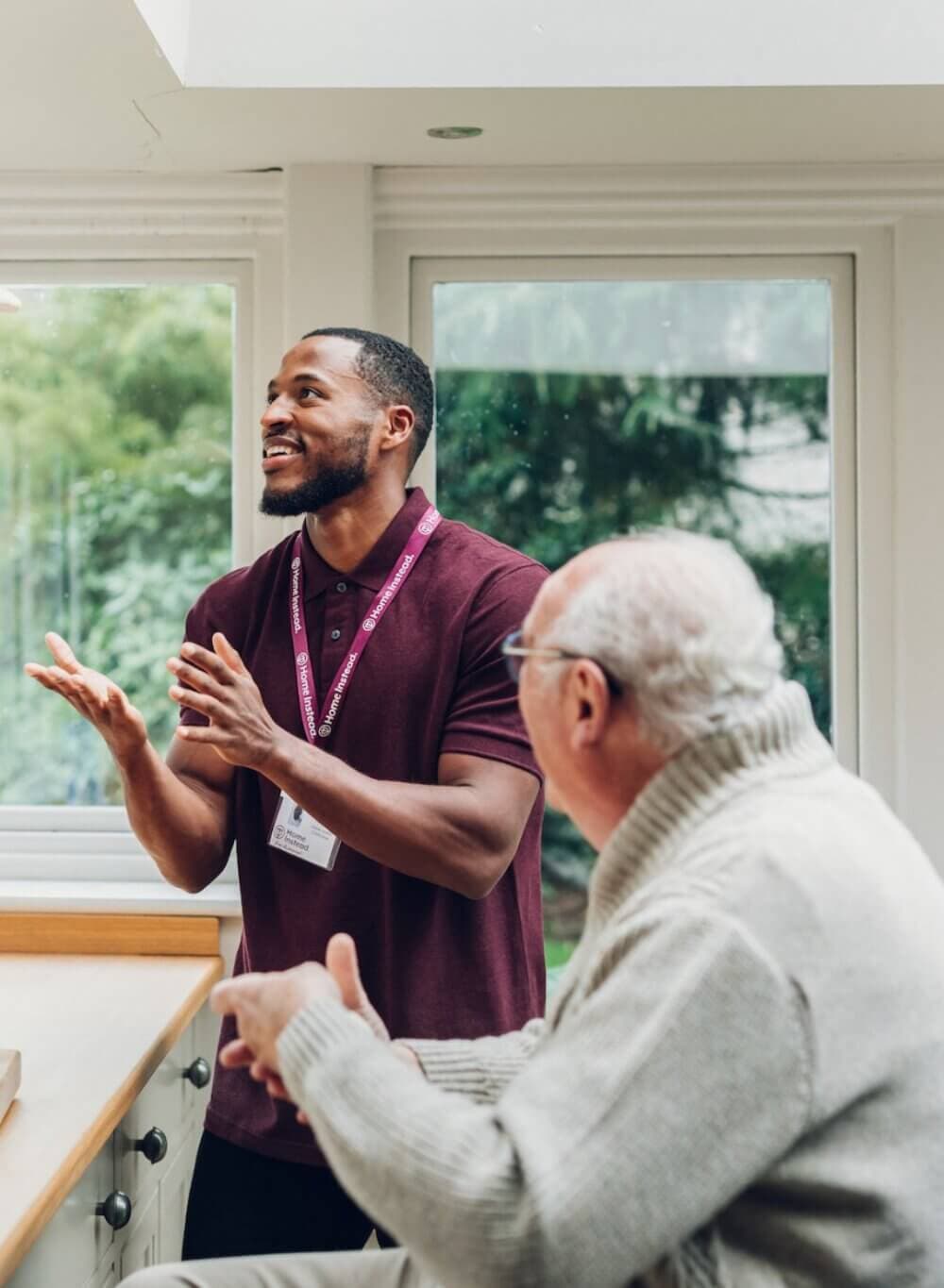 A man in a maroon shirt gestures while talking to an older man in a cream sweater in a bright room. - Home Instead