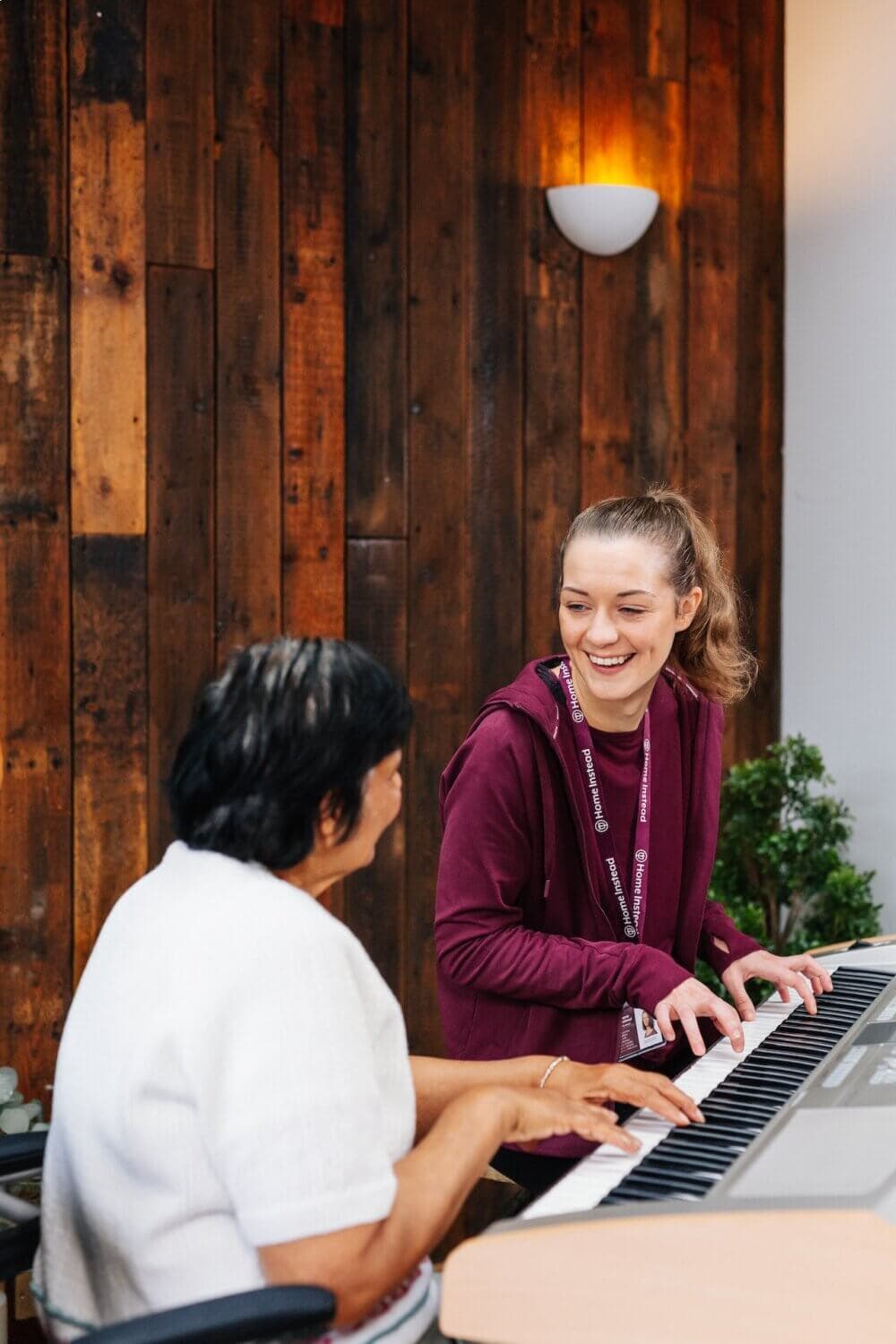 A young woman helps an older woman play the piano, both smiling in a room with wooden walls. - Home Instead