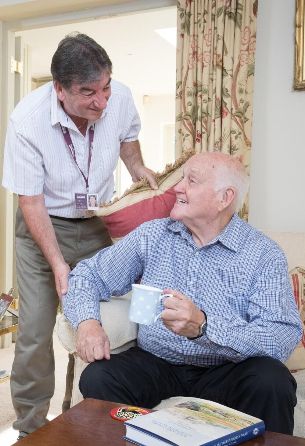 An elderly man with a mug sitting and smiling up at a standing man who is leaning on the couch and also smiling. - Home Instead