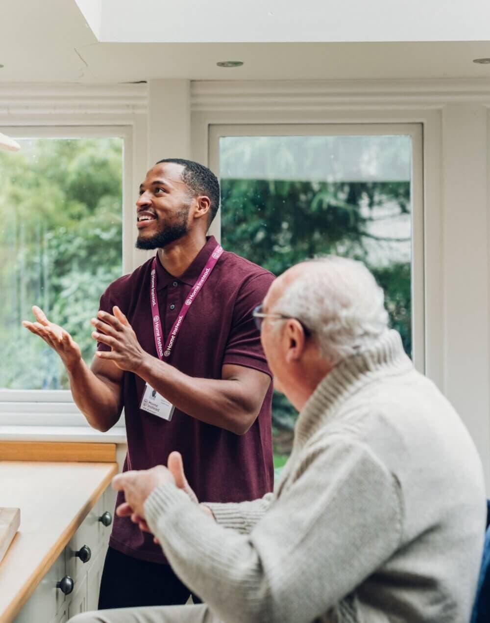 A young man and an older man having a conversation in a bright room with large windows and a wooded view outside. - Home Instead