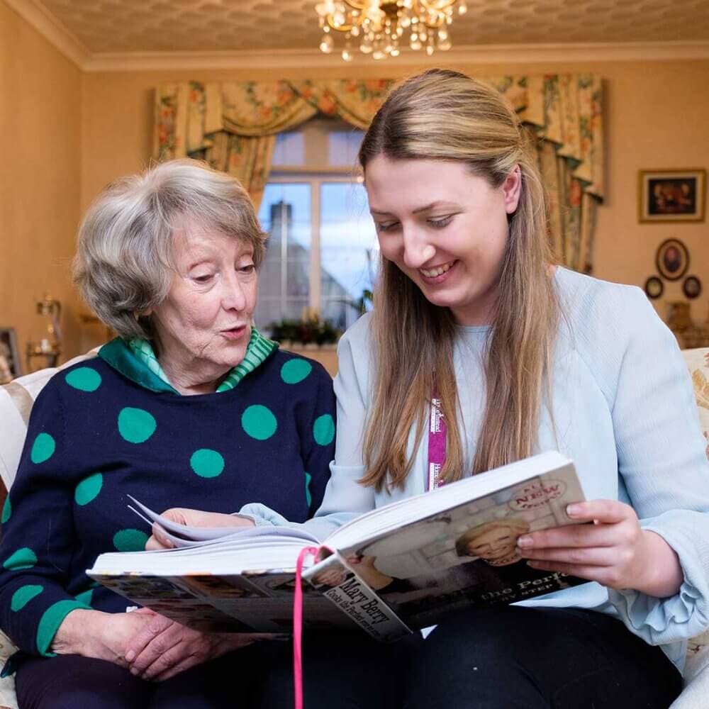 An elderly woman and a young woman sit together on a couch, happily looking through a large book. - Home Instead