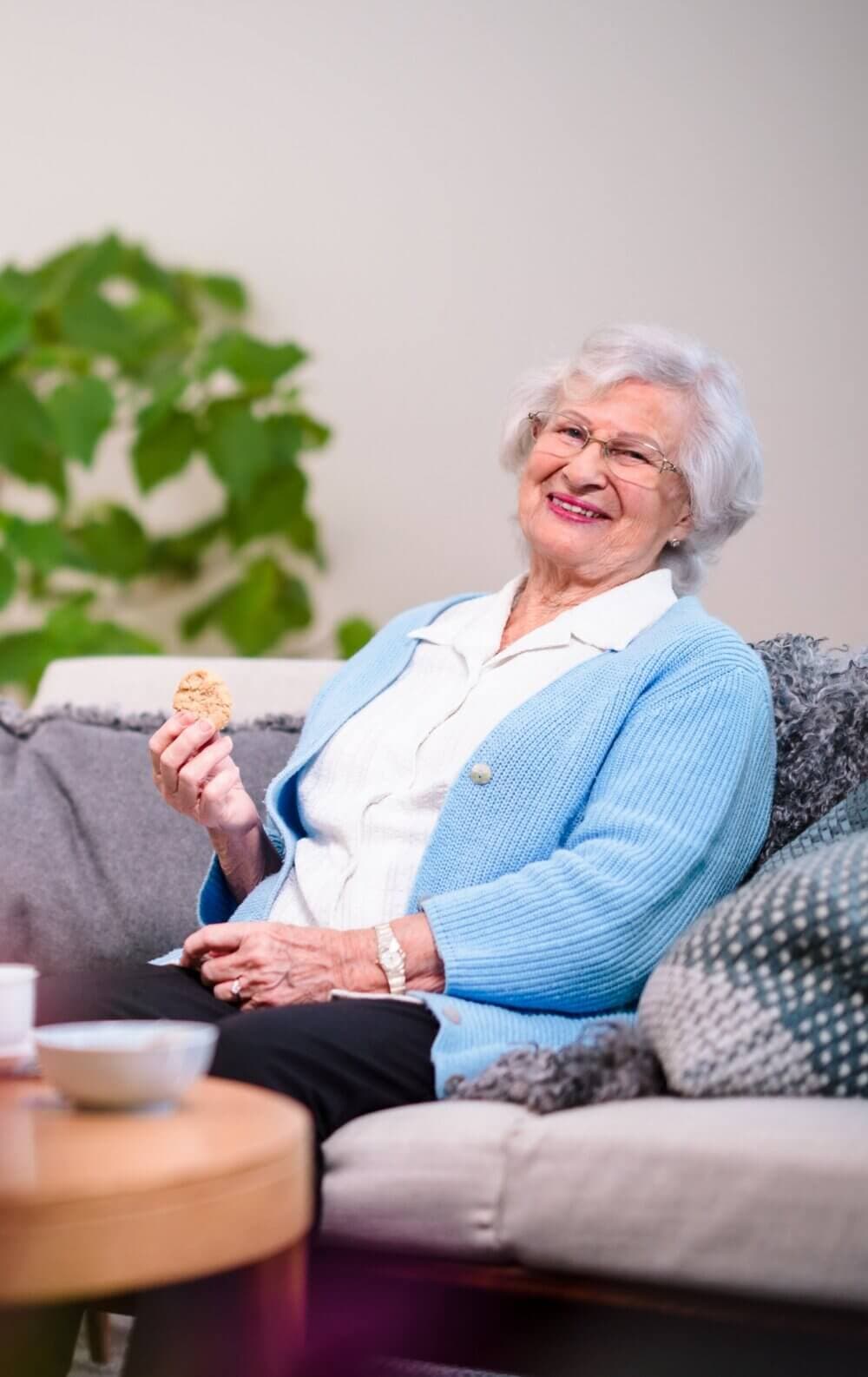Elderly woman in a blue cardigan sitting on a sofa, smiling and holding a snack, with a plant in the background. - Home Instead Bournemouth & Christchurch