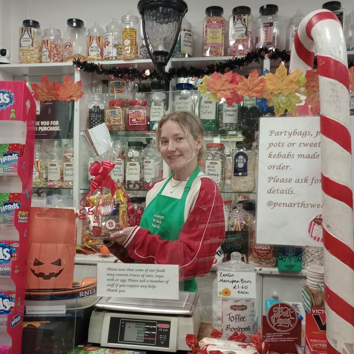 A smiling shopkeeper in a green apron holds a wrapped gift inside a sweets shop decorated for Halloween and autumn. - Home Instead