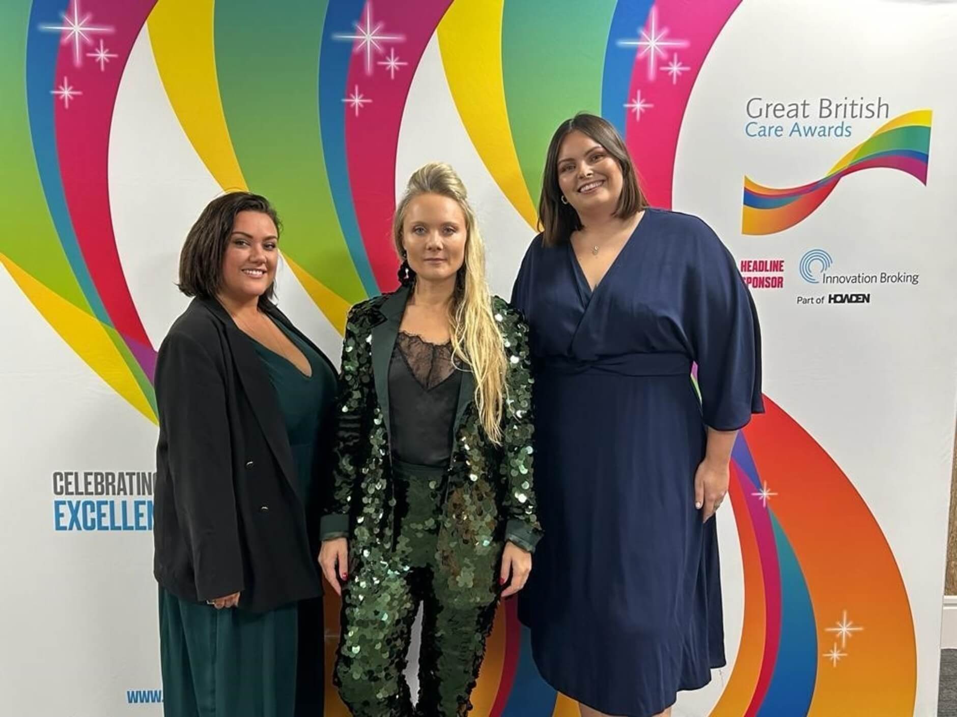 Three women in formal attire standing together at the Great British Care Awards with a colorful backdrop. - Home Instead
