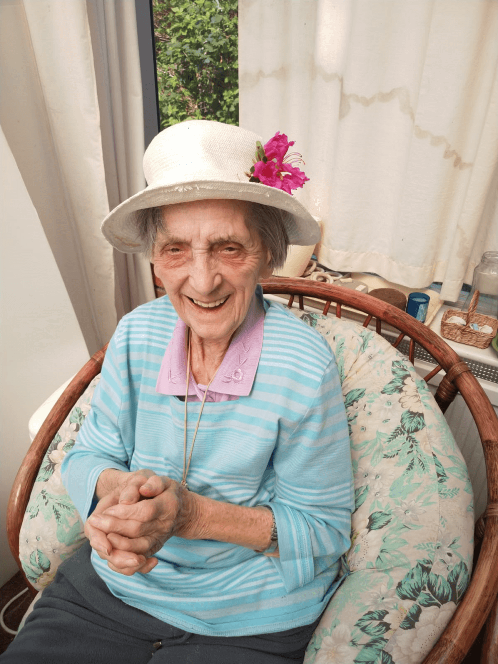 An elderly woman with a hat adorned with a flower sits in a wicker chair, clasping her hands and smiling warmly. - Home Instead