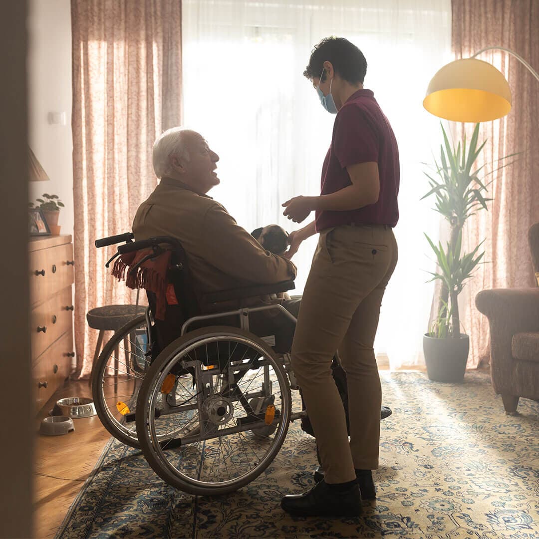 A caregiver wearing a mask assists an elderly person in a wheelchair in a warmly lit room with plants and furniture. - Home Instead