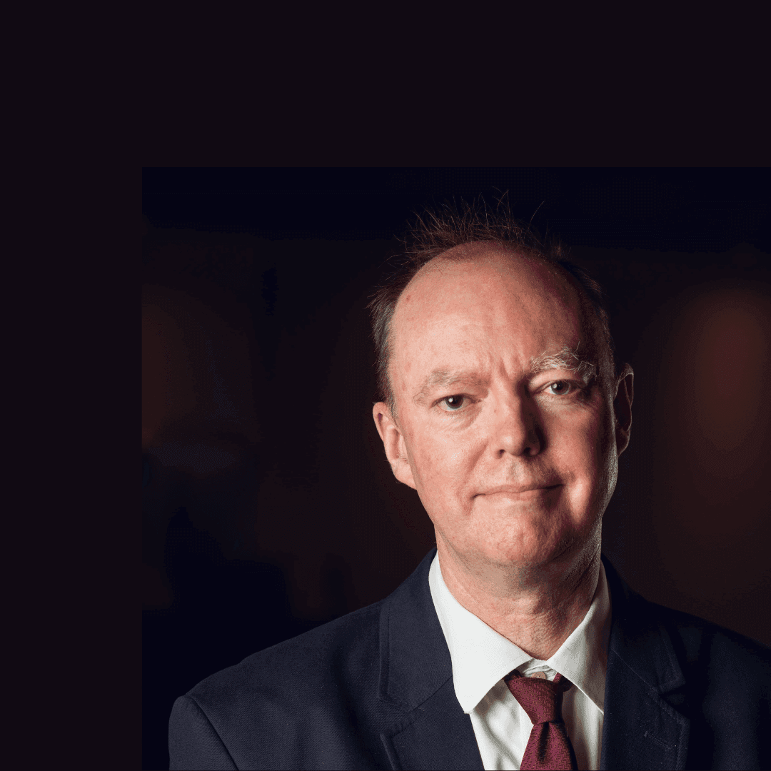 A man in a suit and tie with a neutral expression stands against a dark background. - Home Instead