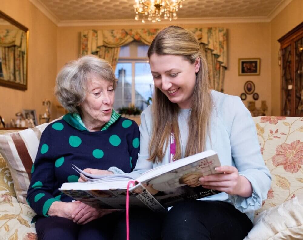 An elderly woman and a young woman smile while looking at a book together on a cozy living room couch. - Home Instead