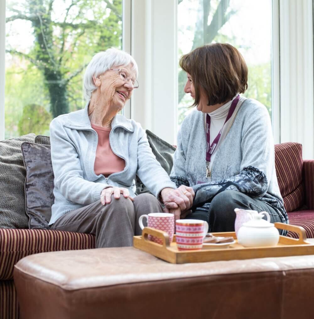 An elderly woman and a younger woman sit on a couch, smiling and holding hands, with tea cups on a tray in front of them. - Home Instead
