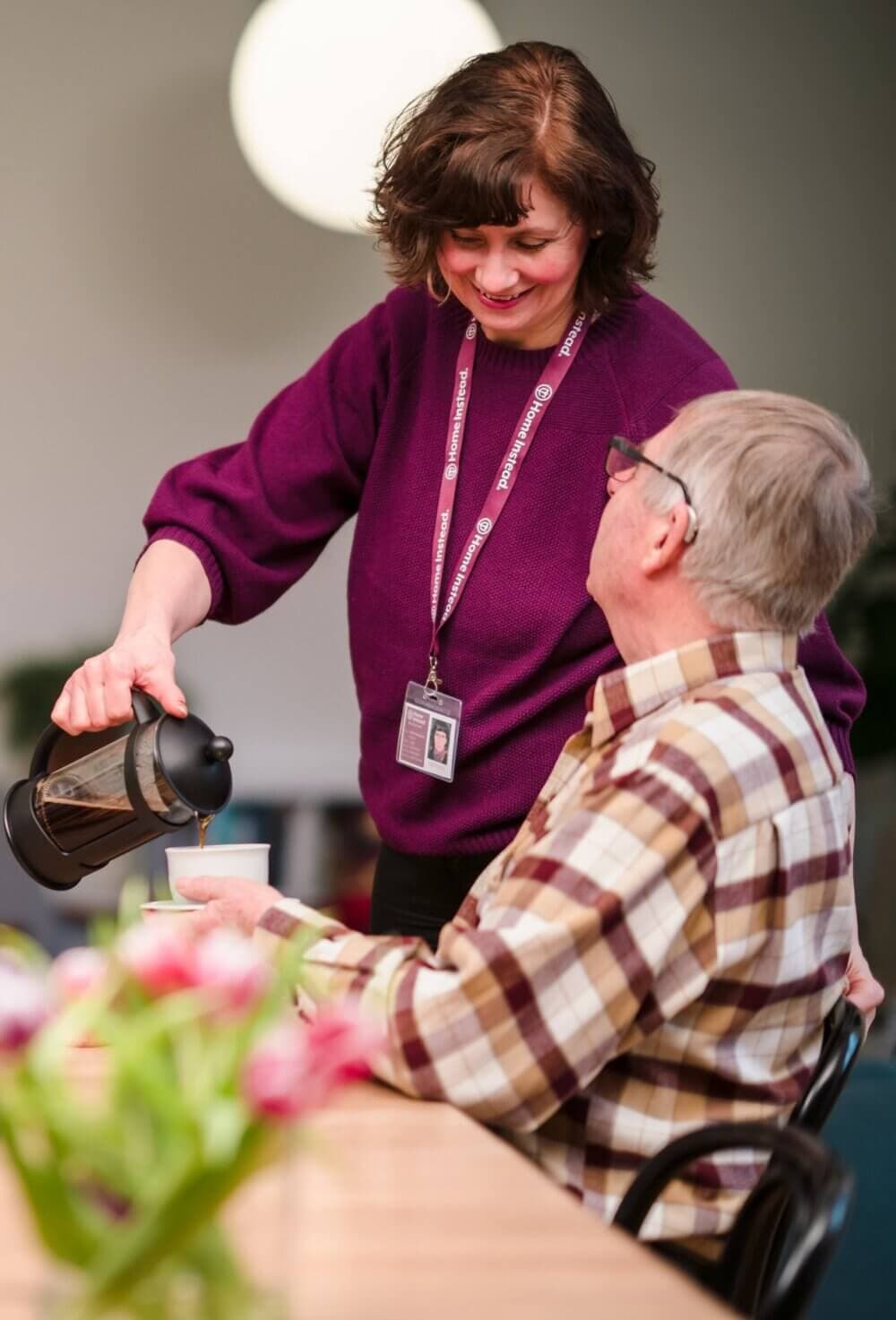 Woman in a purple sweater pours coffee for an elderly man at a table with flowers in the foreground. - Home Instead
