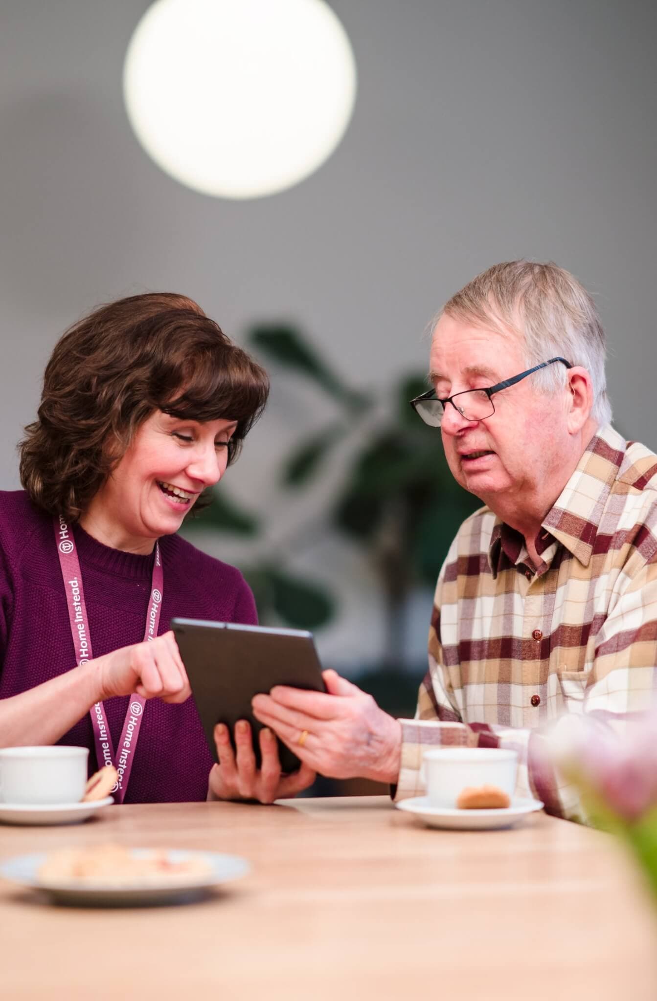 A woman shows a digital tablet to an elderly man at a table with coffee cups. Both are smiling. - Home Instead