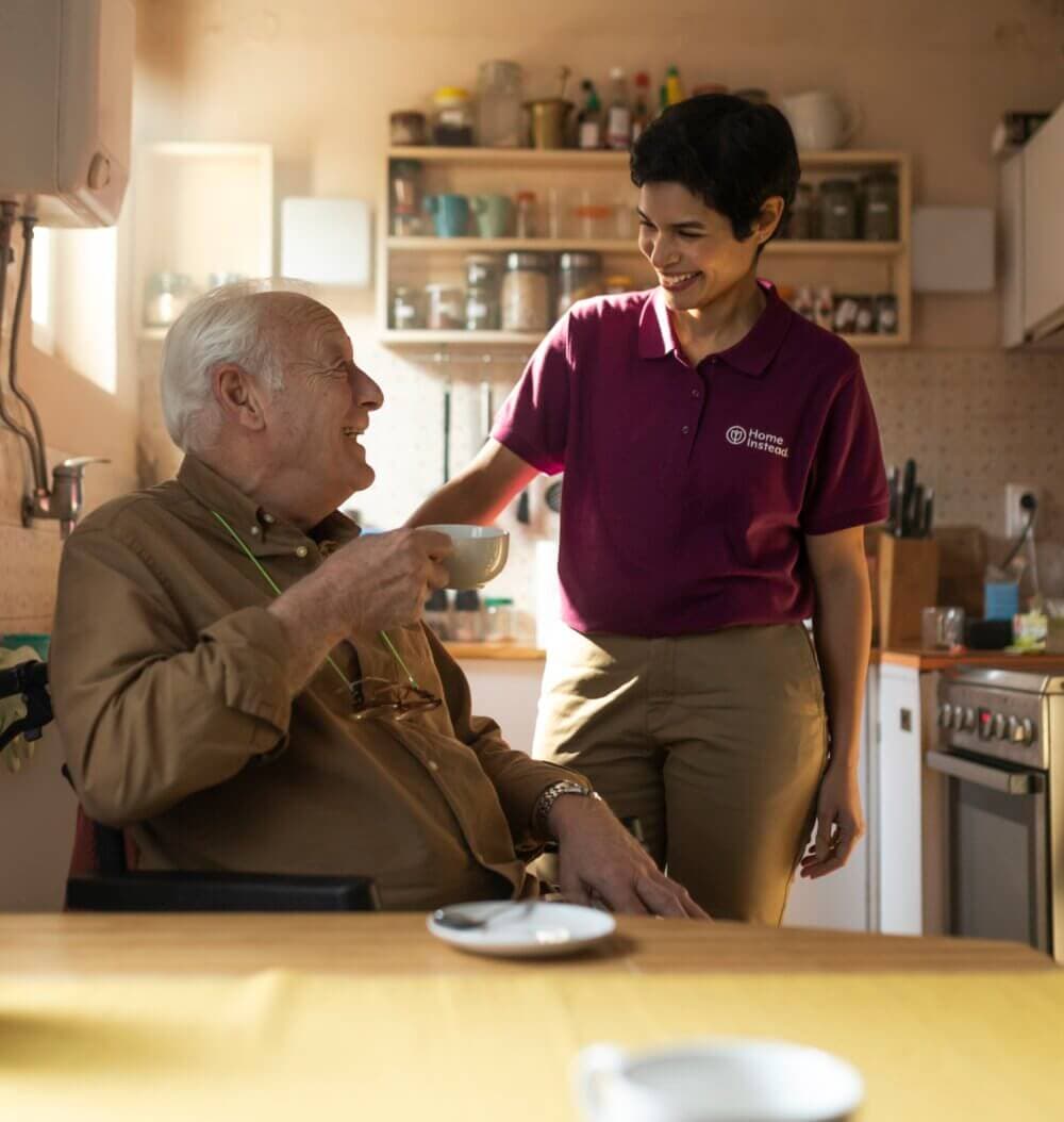 A caregiver stands smiling at an elderly man holding a cup while sitting at a kitchen table. - Home Instead