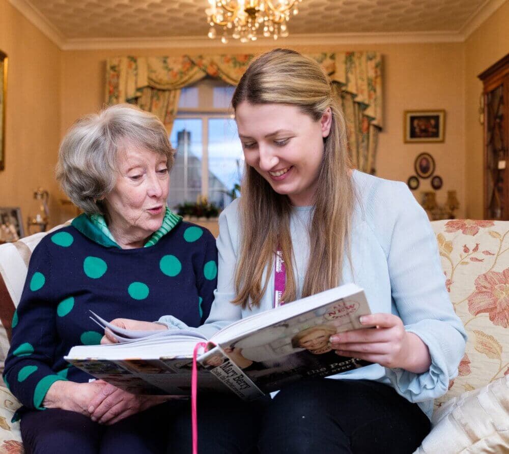 An elderly woman and a young woman sit on a couch, smiling and looking at a magazine together in a cozy living room. - Home Instead