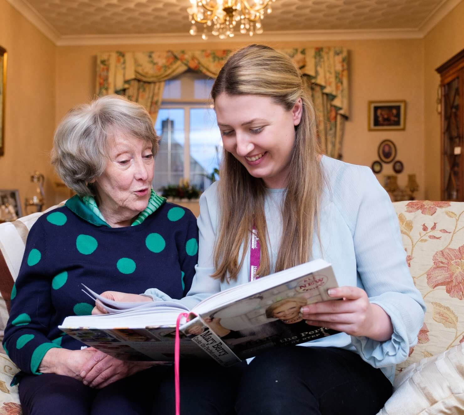 An elderly woman and a young woman sit on a couch, smiling and looking at a magazine together in a cozy living room. - Home Instead