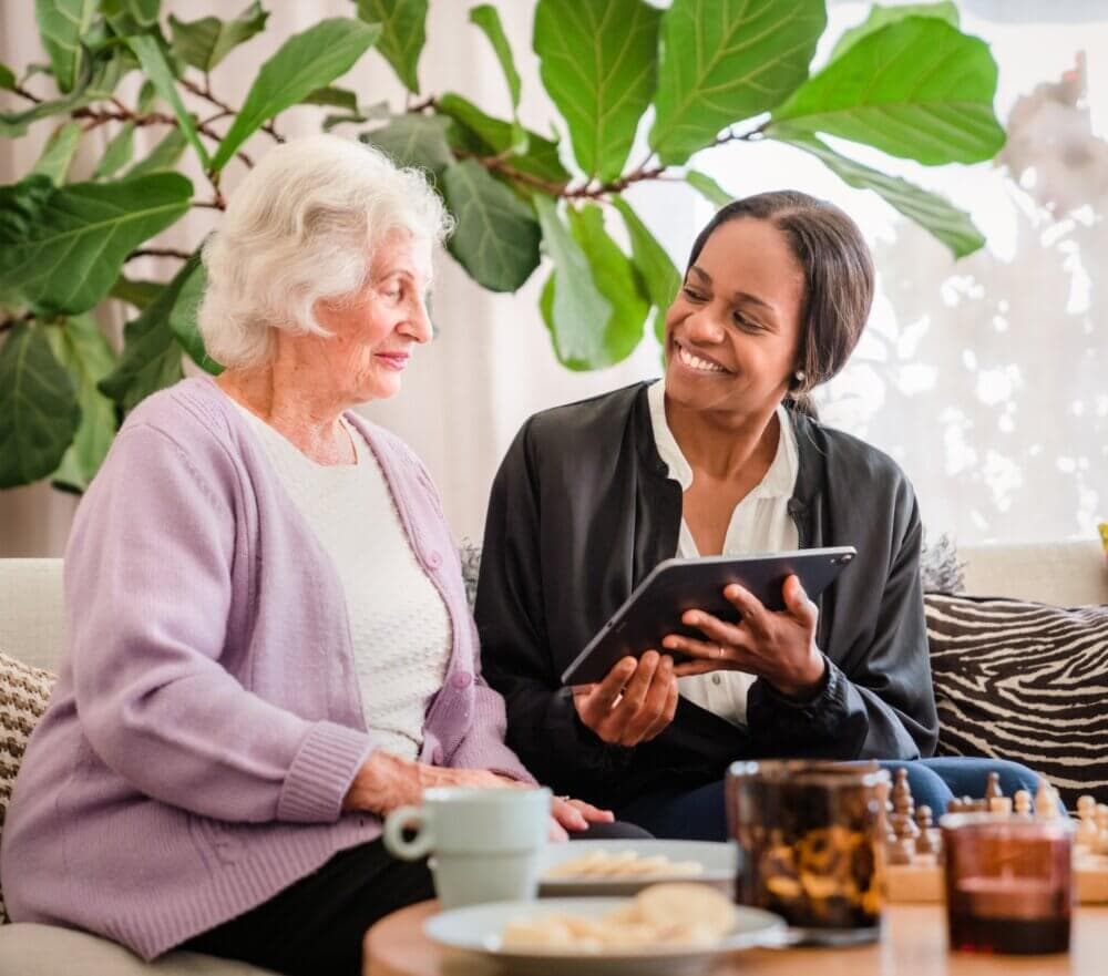 An elderly woman and a younger woman smiling and looking at a tablet together while sitting on a couch. - Home Instead