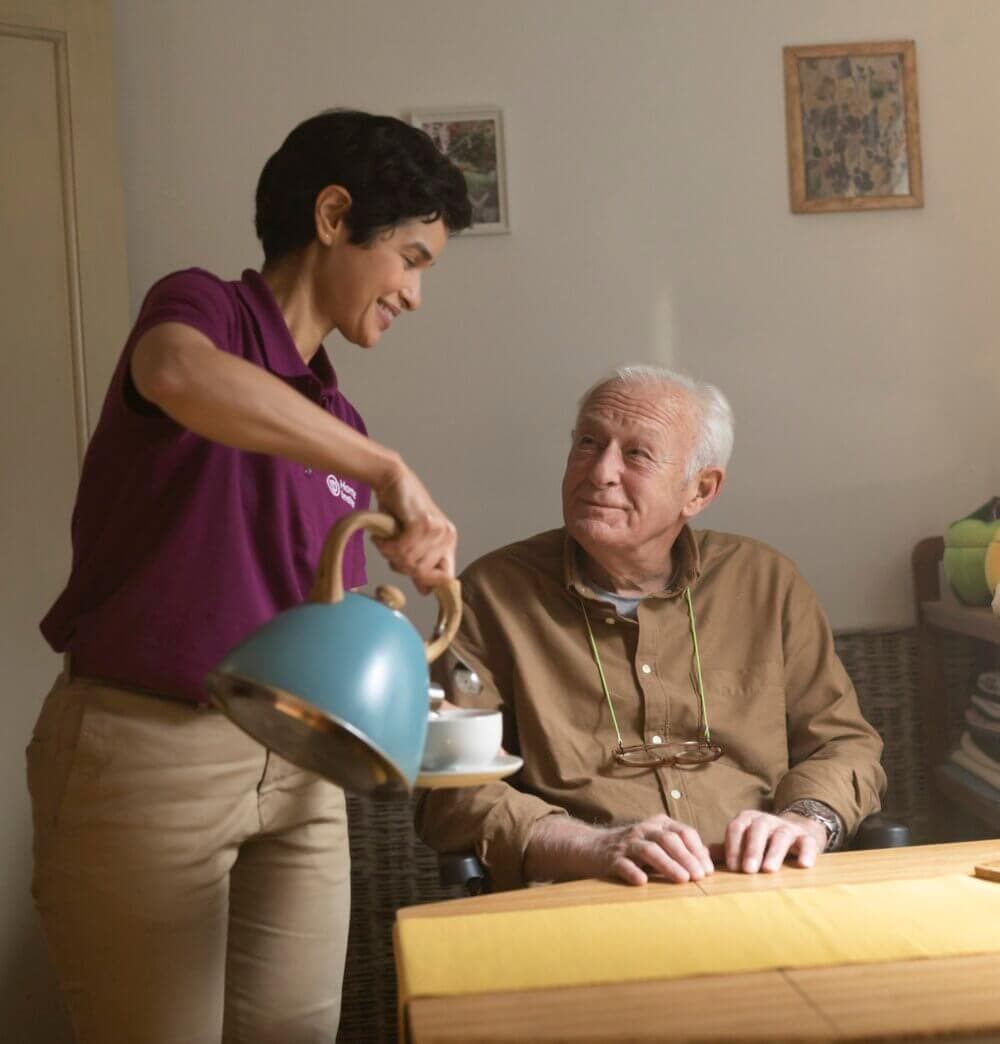 A smiling caregiver in a purple shirt pours tea for a seated elderly man at a table in a cozy room. - Home Instead