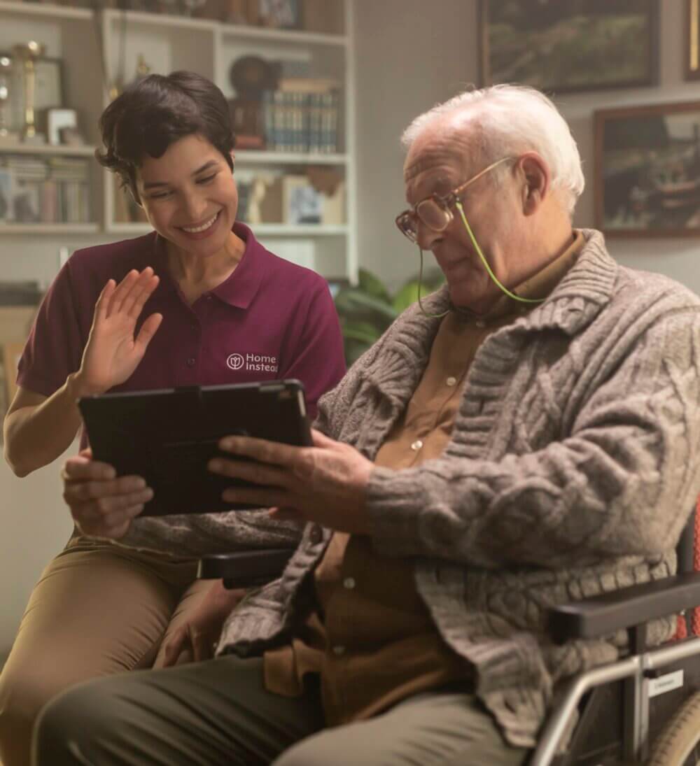 Caregiver smiling and waving at an elderly man in a wheelchair holding a tablet. - Home Instead