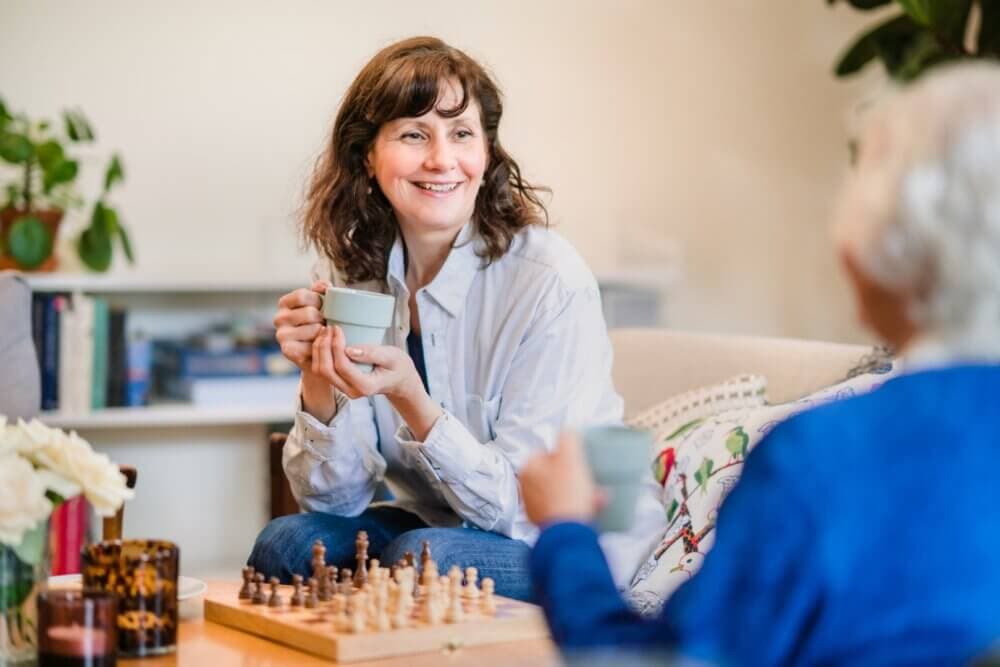 Two people enjoying tea and conversation on a sofa with a chessboard in the foreground. - Home Instead