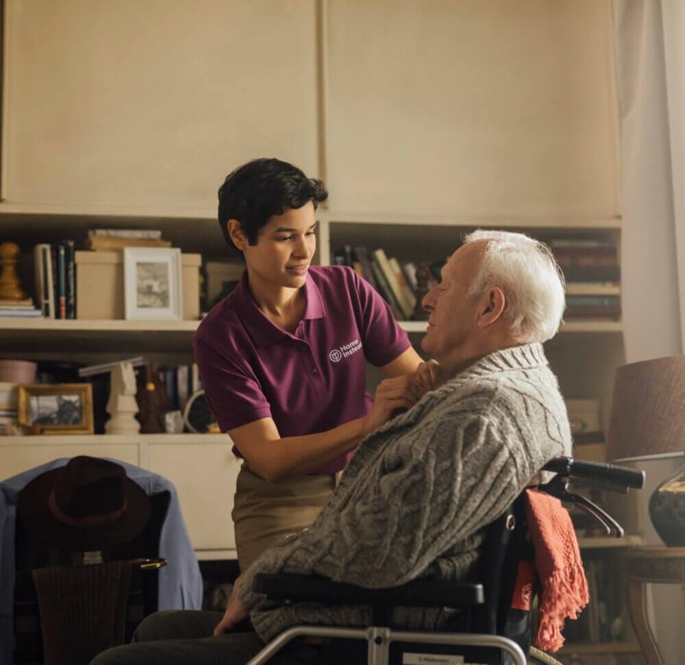 A caregiver helps a wheelchair-bound elderly man in a cozy, book-filled room. - Home Instead